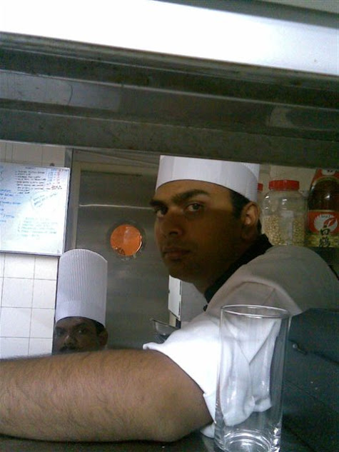 A chef wearing a white hat and uniform looks directly at the camera from behind a counter, with another chef partially visible in the background.