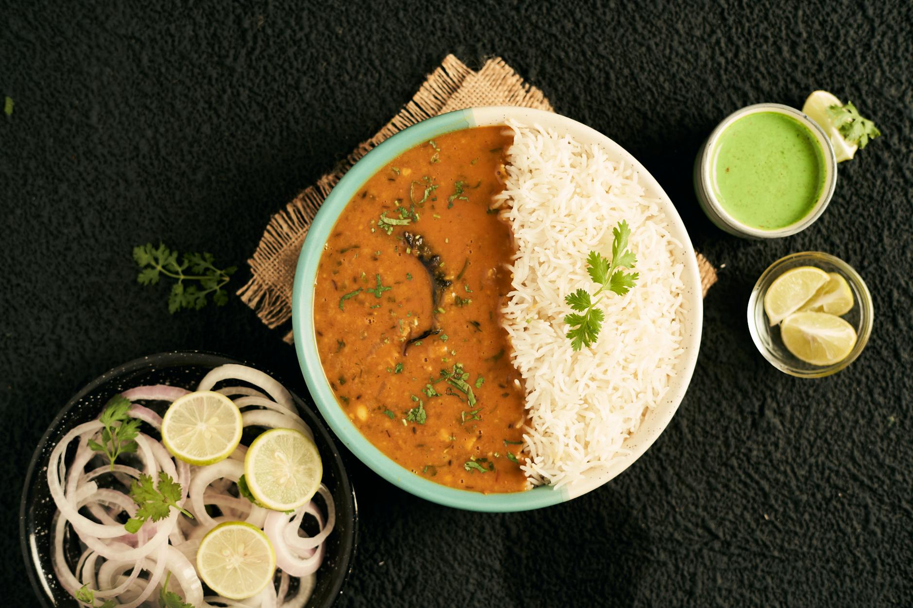 A bowl of rice served with lentil curry, garnished with fresh coriander. Accompanied by a small bowl of green chutney, lemon wedges, and sliced onions on the side.