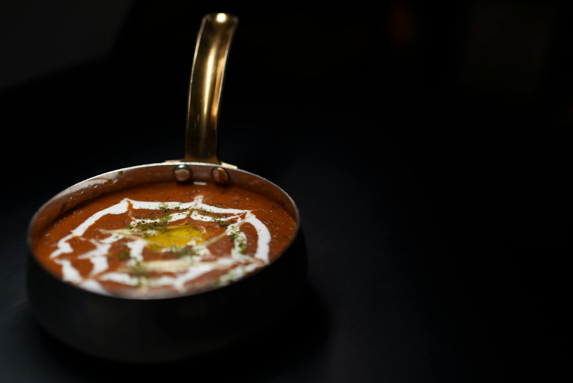 A close-up of a bowl of creamy curry garnished with swirls of yogurt and fresh herbs, set against a dark background.