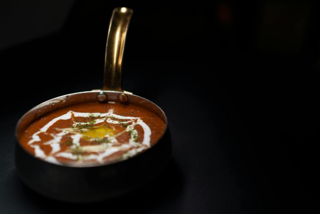 A copper bowl filled with a rich, red curry topped with a swirl of cream and garnished with herbs, set against a dark background.