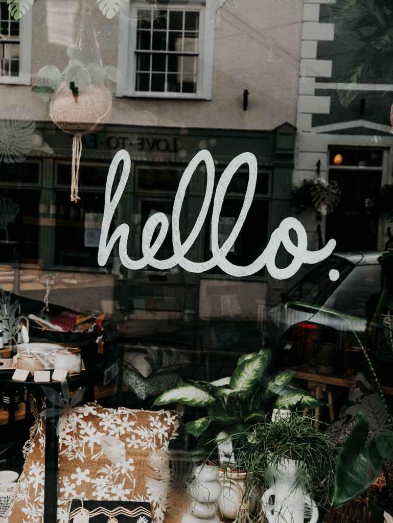 A close-up of a store window featuring the word 'hello.' in a playful, handwritten style. The background shows various decorative items like cushions and plants.